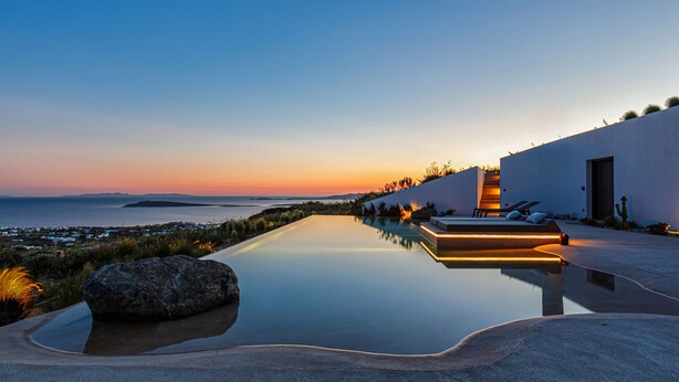 A modern infinity pool with a view of the sea at sunset, a white building with a staircase, and outdoor seating. - Paros Villa Skylines Estate I (Paros)
