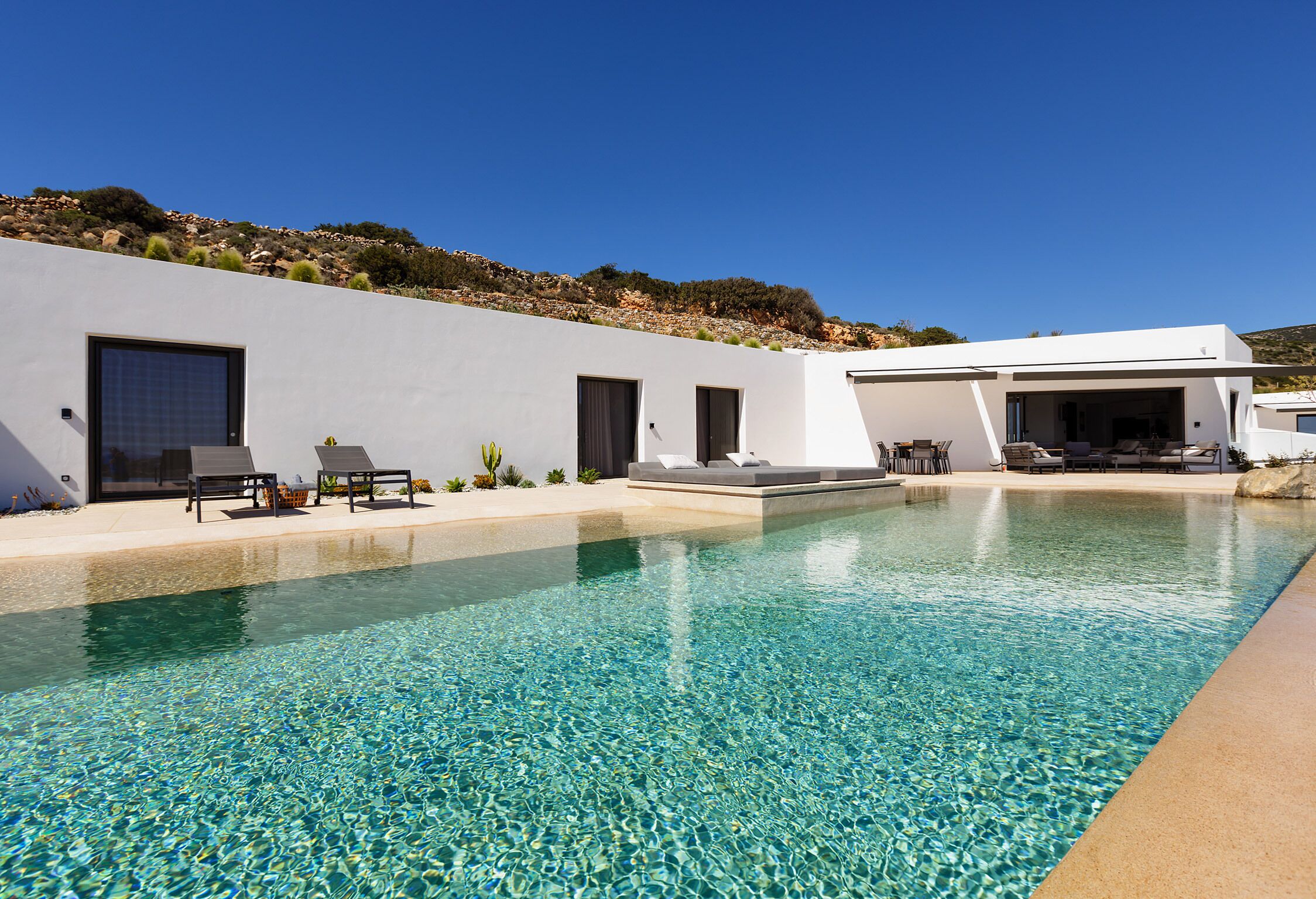 Un espace piscine moderne avec une eau cristalline, des chaises longues et un bâtiment blanc doté de grandes fenêtres et de grandes portes.