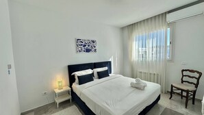 Ein ordentlich eingerichtetes Hotelzimmer mit einem großen Bett, einem Nachttisch mit Lampe, einem Sessel und einem Fenster mit Vorhängen.
