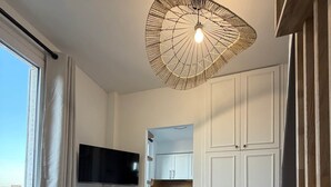 Ett modernt vardagsrum med en platt-TV, ett runt bord med en vävd bordsduk, en unik taklampa och utsikt över köket genom en öppen dörr.