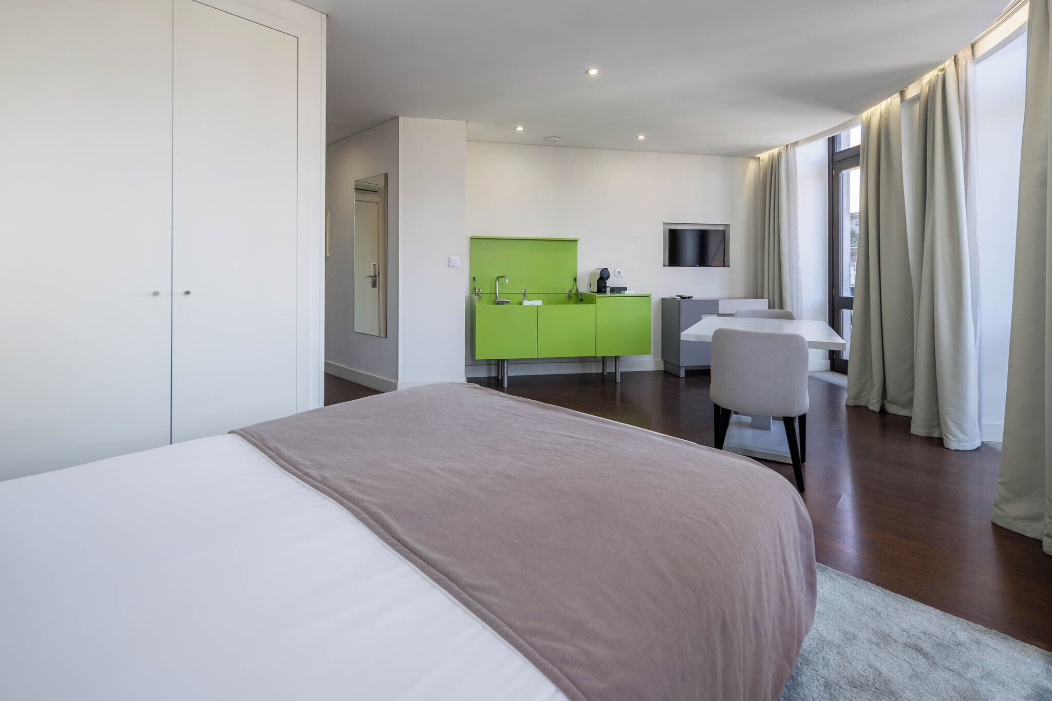 Habitación de hotel con cama, escritorio, minibar verde, televisión y un amplio ventanal con cortinas.