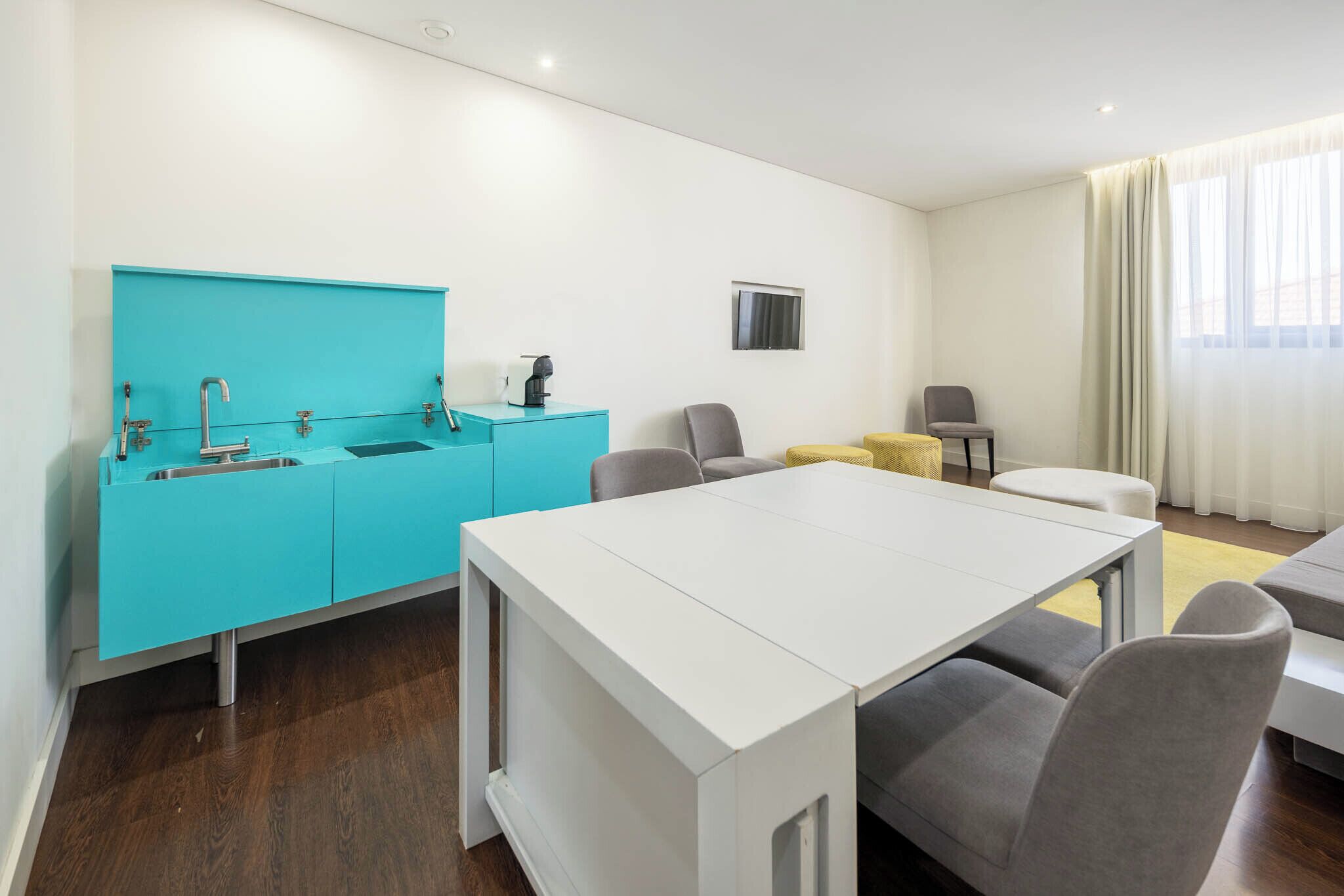 A modern hotel room with a white dining table, grey chairs, a yellow ottoman, a turquoise kitchenette, and a large window with curtains.