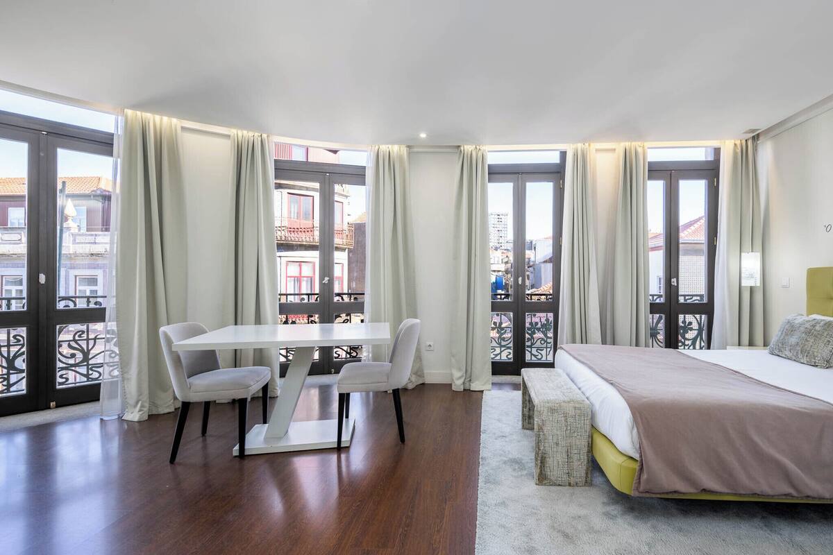 Una habitación de hotel con cama, mesa de comedor con sillas, un banco y ventanas grandes con cortinas.