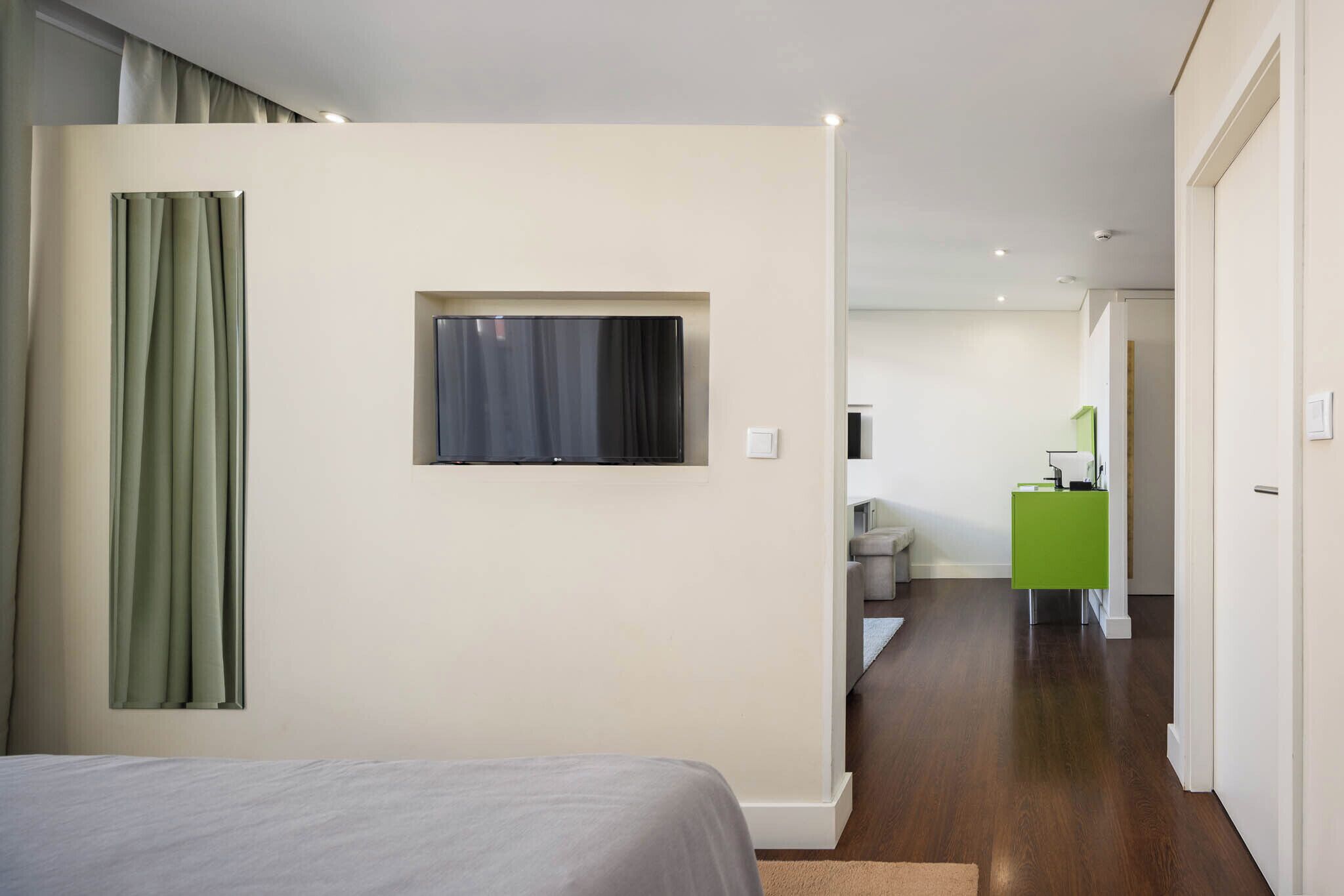 A modern hotel room with a bed, a TV mounted on the wall, a green desk, and a hallway leading to a sitting area.