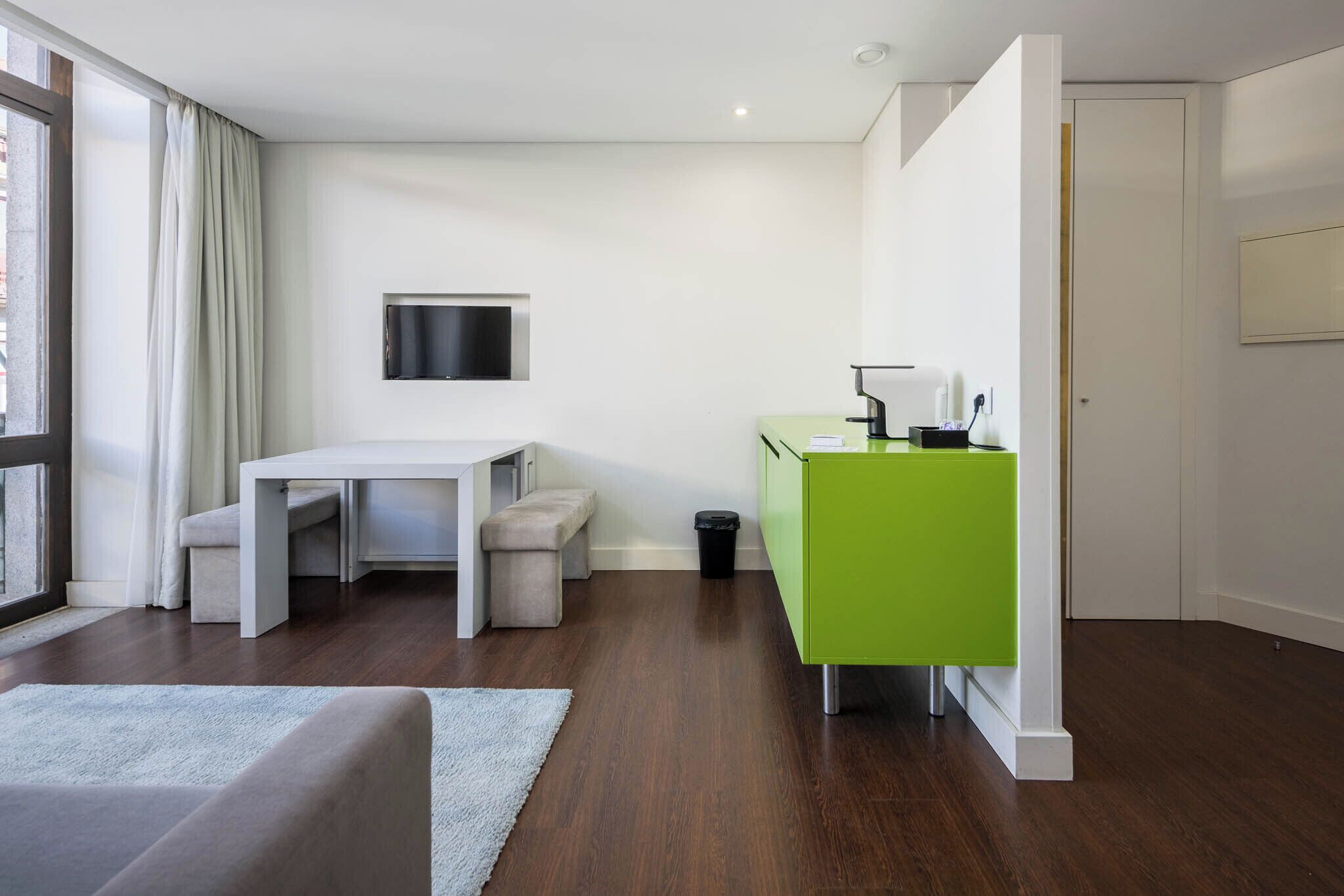Una habitación moderna con un escritorio blanco, un banco y un armario verde, un televisor montado en la pared y una ventana con cortinas.