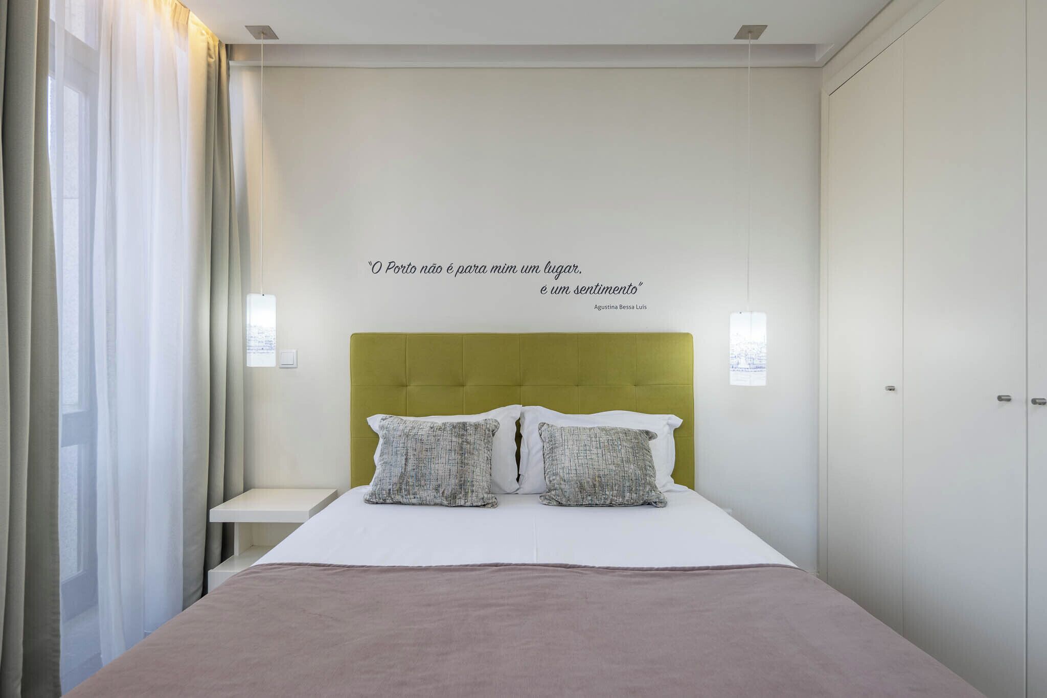 A hotel room with a bed, a bedside table, a wall quote, and a window with curtains.