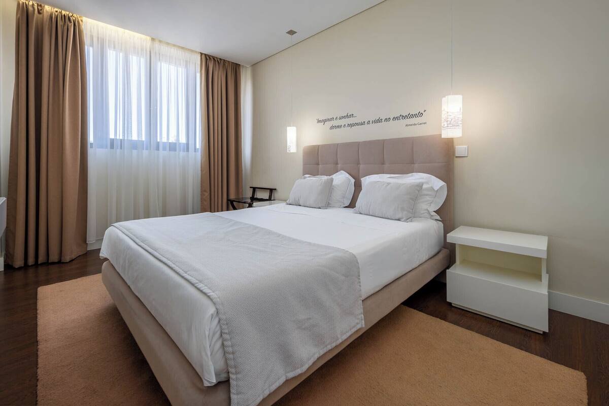 Una habitación de hotel moderna con una cama grande, una mesita de noche, una lámpara de pared y una ventana con cortinas.