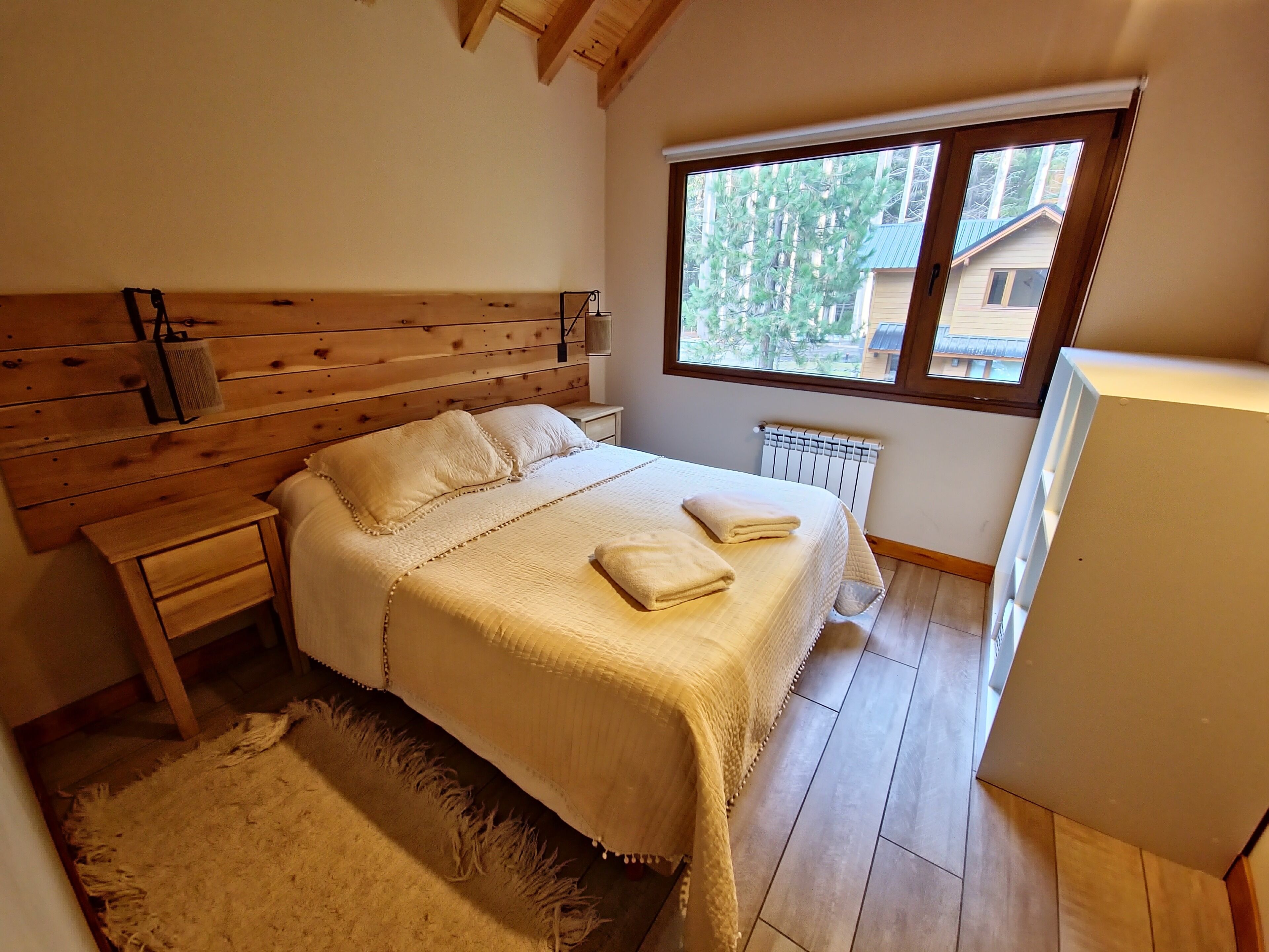 A bedroom with a wooden headboard, a large window, a radiator, and a white bedspread.