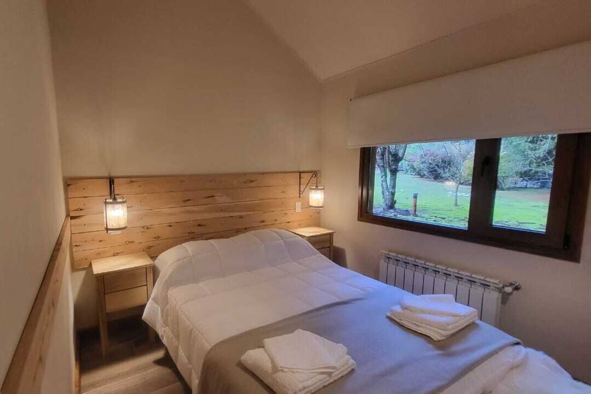 Een slaapkamer met een houten hoofdbord, een bed met witte beddengoed, twee nachtlampjes, een raam met uitzicht op groen, en een hangend horloge.