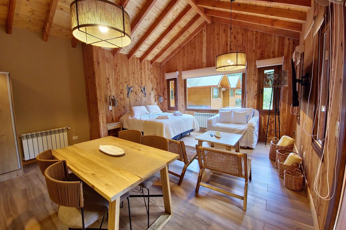 Een gezellig houten kamer in chaletstijl, met een eethoek met tafel en stoelen, een bed met een wit dekbed en een groot raam dat natuurlijk licht binnenlaat.