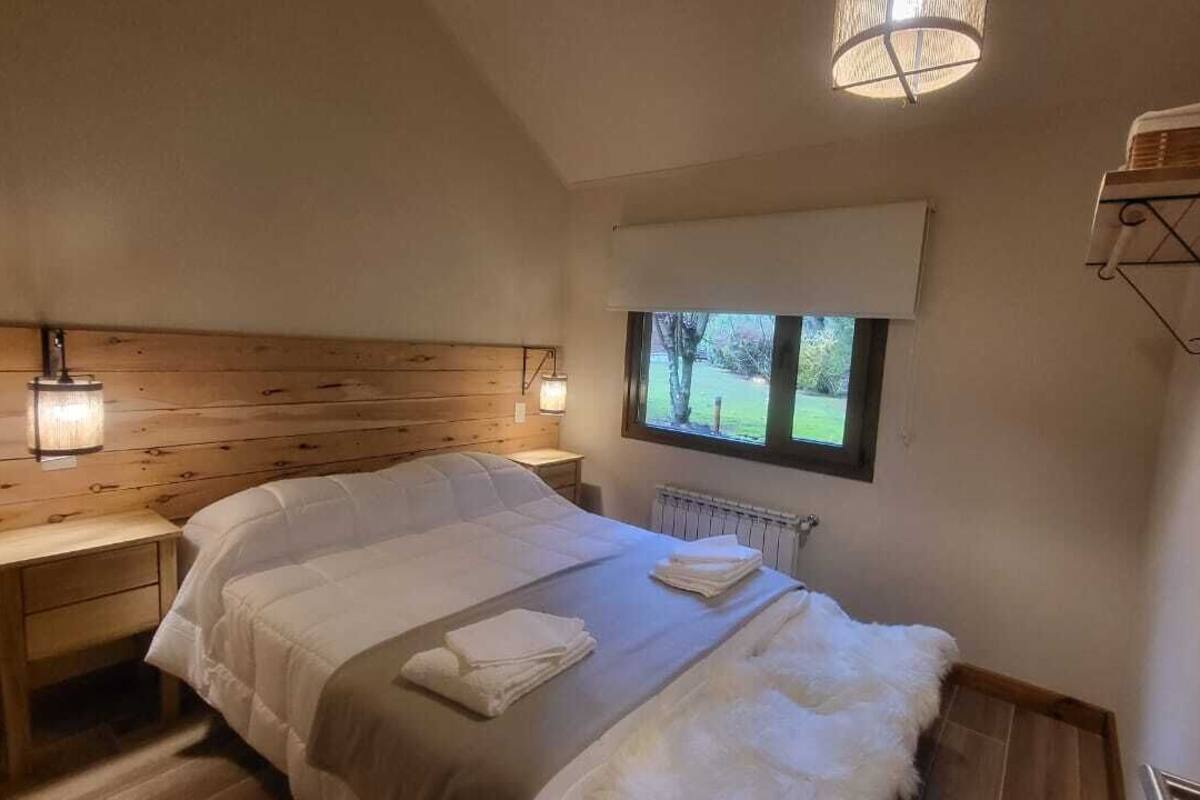Een slaapkamer met een houten hoofdeinde, een groot bed met witte beddengoed, een raam met uitzicht op groen, en een wandplank met handdoeken.