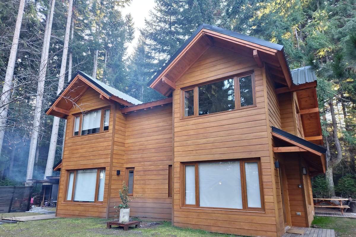 Een houten chalet met twee verdiepingen, grote ramen en een terras, omringd door hoge bomen.
