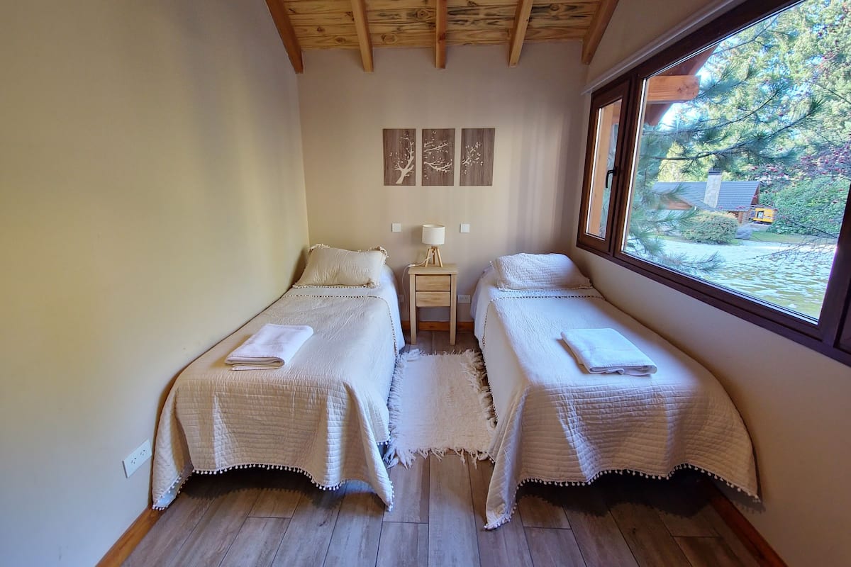 Een kleine, gezellige kamer met twee bedden, een nachtkastje, een lamp en een raam met uitzicht op bomen en een gebouw.