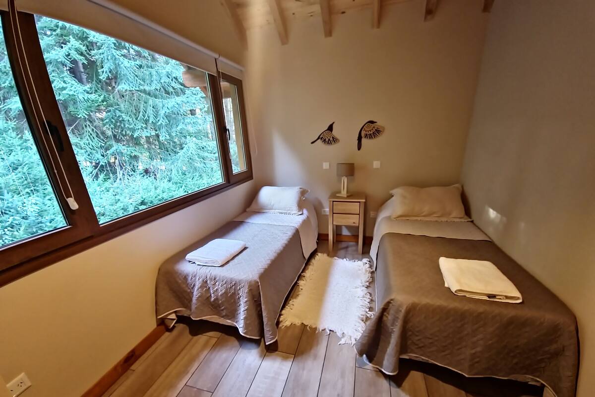 Een slaapkamer met twee bedden, een nachtkastje, een lamp en een raam met uitzicht op bomen.