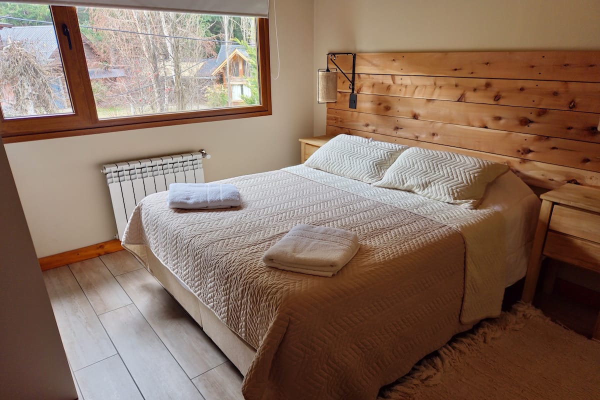Een slaapkamer met een houten hoofdbord, een groot raam, een radiator en een bed met een gewatteerde spreideken en kussens.