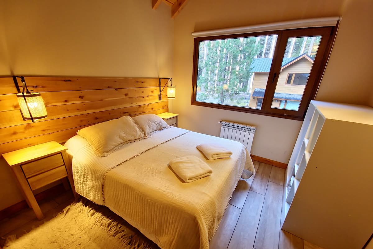 Een slaapkamer met een houten hoofdbord, een bed met witte beddengoed, twee bedlampjes, een raam met uitzicht op bomen, en een radiator.