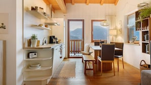 Cocina compacta con estantes de madera, comedor con banco y vistas a montañas a través de un ventanal.