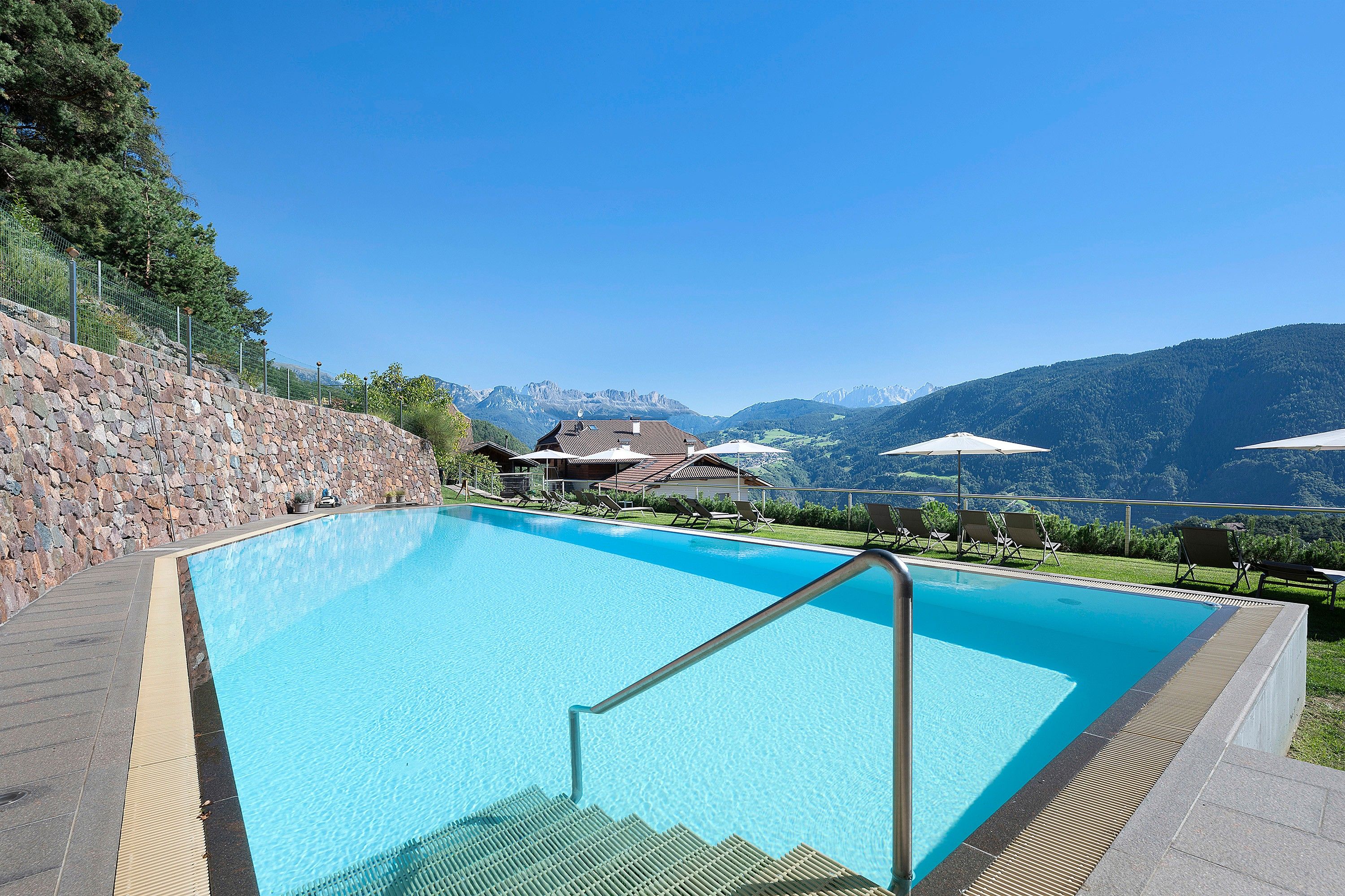 Ein klares, blaues Schwimmbecken mit einer Steinmauer an einer Seite, Liegestühlen und Blick auf Berge im Hintergrund.
