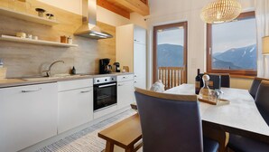 Una cocina moderna con vigas de madera, un área de comedor con mesa y sillas, y vistas a montañas a través de grandes ventanales.