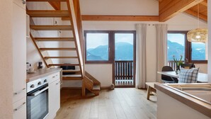 Una cocina con vigas de madera, una escalera, vistas a las montañas, un comedor con mesa y sillas, y un amplio ventanal con cortinas.