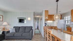 Ein gemütliches Wohnzimmer mit grauem Sofa, Fernseher und Lampe. Es gibt einen Essbereich mit Holzsesseln und Tisch. Die Küche verfügt über Holzschränke und eine Pendelleuchte.