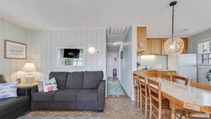 A modern living room with a grey sofa, a dining table with wooden chairs, a kitchen area with wooden cabinets, and a hanging pendant light. - AJ: 3 Bedroom Oceanfront Duplex (Carolina Beach)