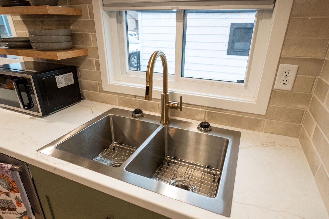 Una cocina con dos fregaderos de acero inoxidable, un microondas negro y una ventana sobre el fregadero.