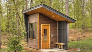 Een klein houten huisje met een houten deur, een bankje ervoor en een omliggend bos.