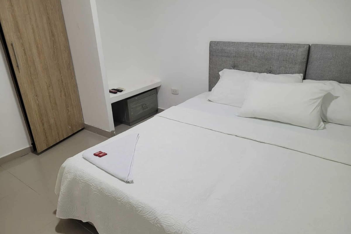 A neatly made bed with white bedding and pillows, a grey headboard, and a small bedside table with a drawer.