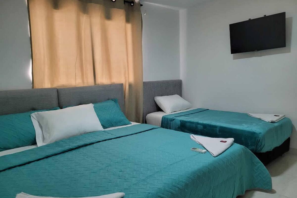 A hotel room with two beds, each with teal bedding and white pillows. There is a flat-screen TV mounted on the wall above the beds.