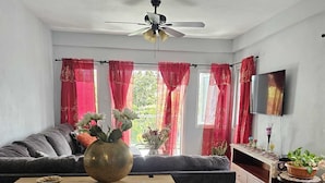 A modern living room with a grey sofa, a wooden coffee table, a ceiling fan, red curtains, a TV, and a potted plant. - House of the Three Companions (Gros Islet)