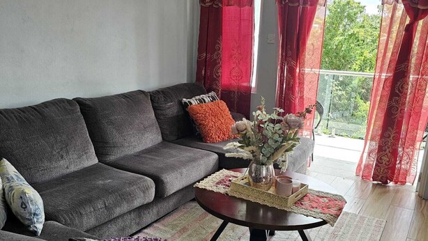 A living room with a grey sofa, a small round table with a vase and a tray, and red curtains. - House of the Three Companions (Gros Islet)