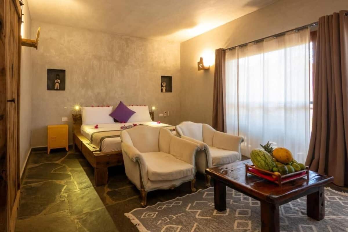 Camera d'albergo con un letto, due poltrone, un tavolino con della frutta e un tappeto decorato.