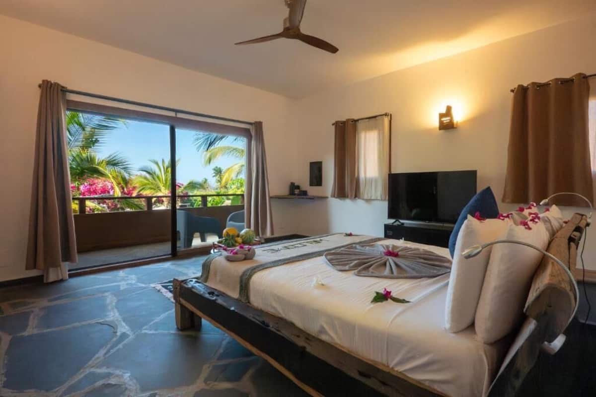Camera d'albergo con un letto, una televisione, una ventola a soffitto e un balcone con vista su palme e fiori.