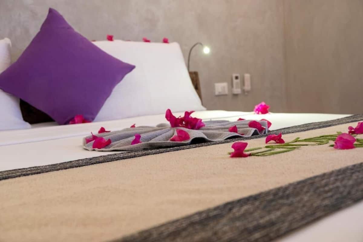 Un letto rifatto con un cuscino bianco e viola, una coperta beige e petali di rosa rosa sparsi.