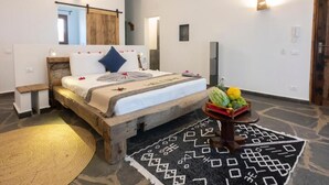 A bedroom with a wooden bed, a bedside table, a wall-mounted light, a mirror, and a fruit basket on the floor. - Di Milo Boutique Hotel (Kiwengwa)