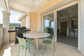 Een terras buiten met een ronde tafel en stoelen, een barbecue en een schuifpui naar een binnenruimte.
