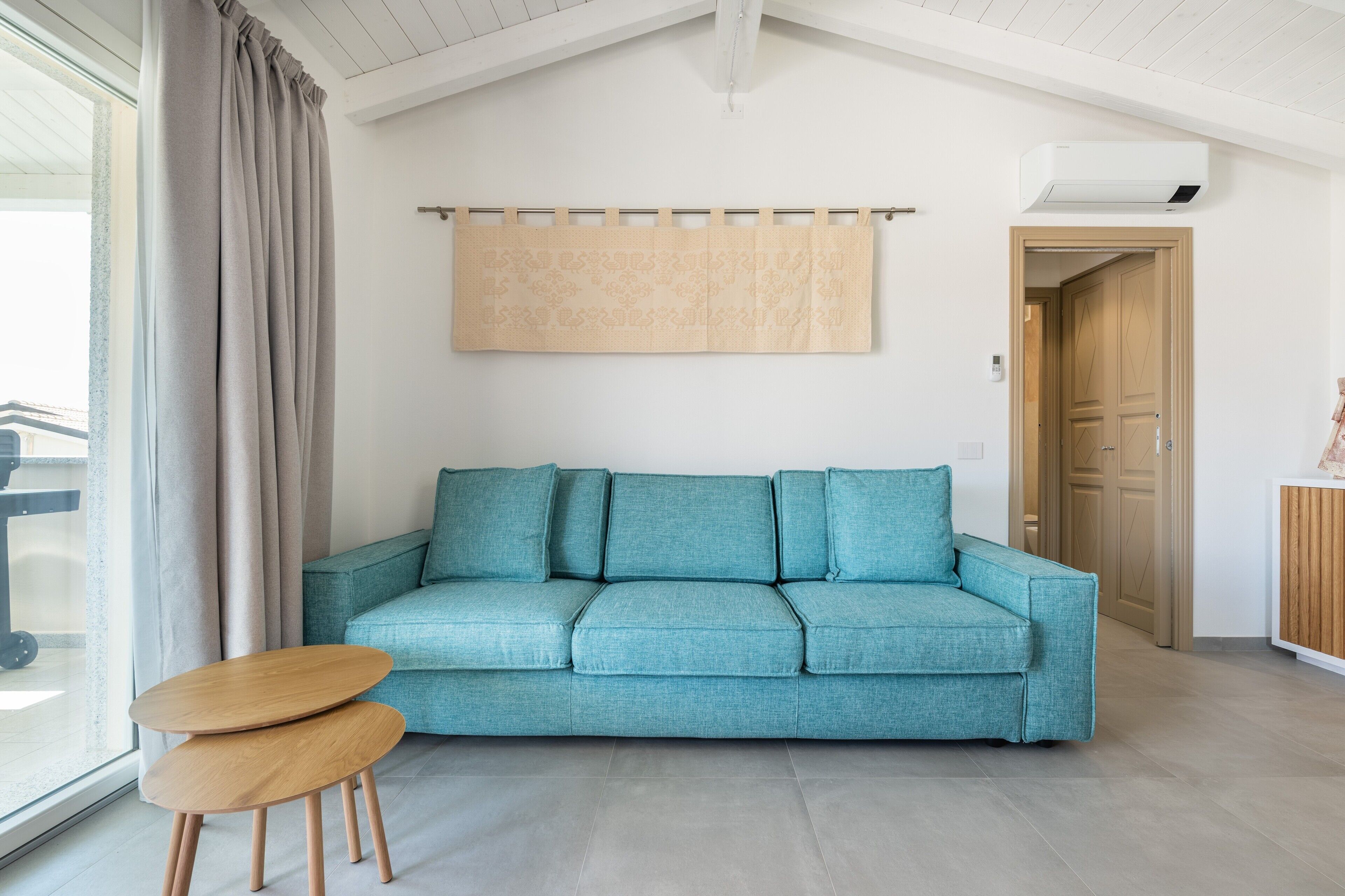 Un sofá color turquesa, dos taburetes de madera, un aire acondicionado instalado en la pared y una puerta de madera.