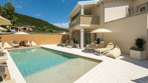 A poolside area with lounge chairs and umbrellas, a pool, and a building with a stone facade and balconies. - Holiday Home 'Borgo Myricae - Calendula' with Sea View, Wi-Fi and Air Conditioning (Chia)