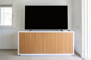 A modern room with a white wall, a wooden panel cabinet, and a large flat-screen TV mounted above it. - Holiday Home 'Borgo Myricae - Scilla Bianca' with Shared Pool, Wi-Fi and Air Conditioning (Chia)