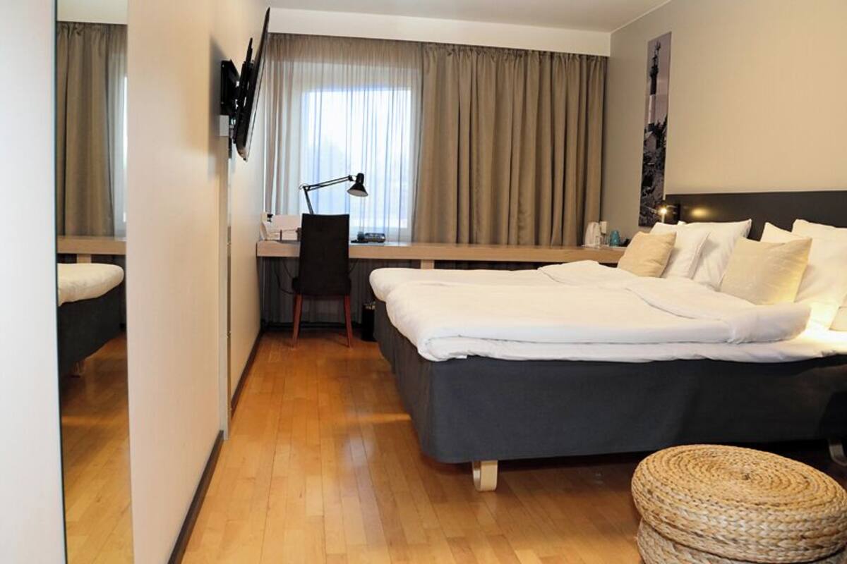 Una camera d'albergo con un letto, una scrivania, una sedia, una lampada e un pouf rotondo.