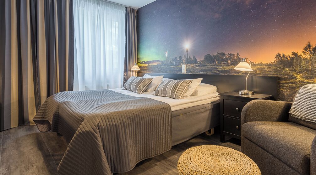 A hotel room with a bed, nightstand, lamp, chair, and a mural of a night sky.
