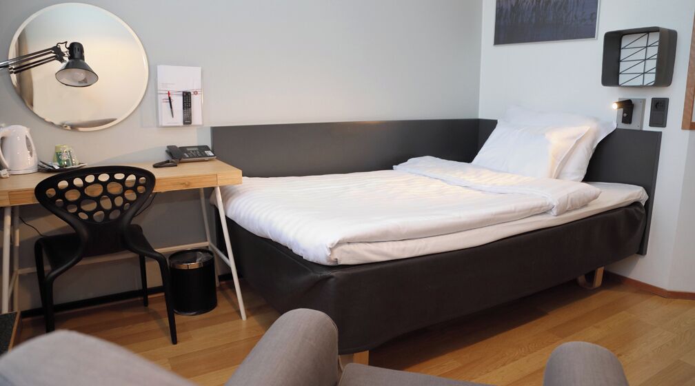 A bedroom with a bed, desk, chair, and a wall-mounted lamp.