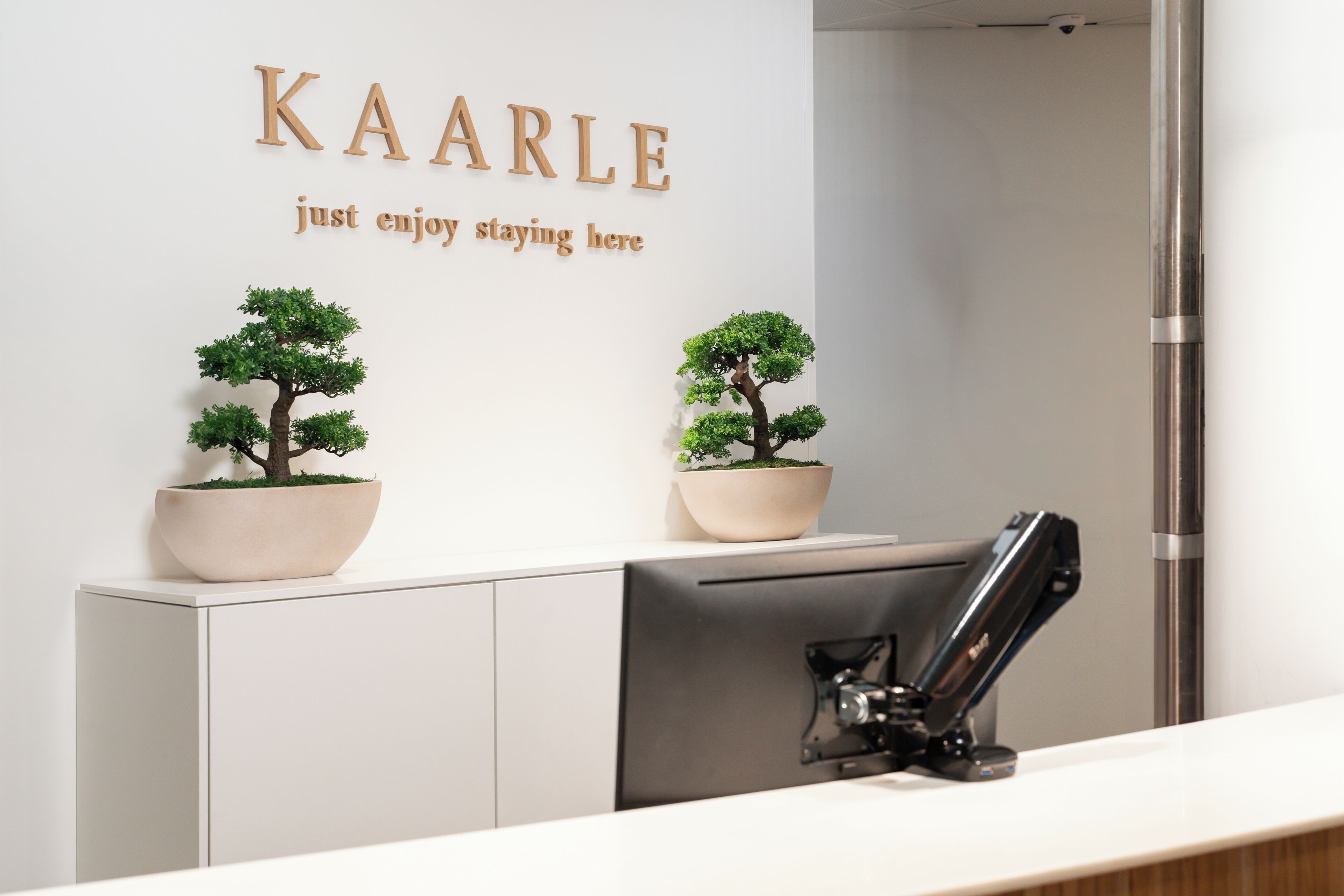 Een receptiebalie met twee potplanten, een bord waarop staat 'KAARLE geniet gewoon van je verblijf' en een beveiligingscamera erboven.