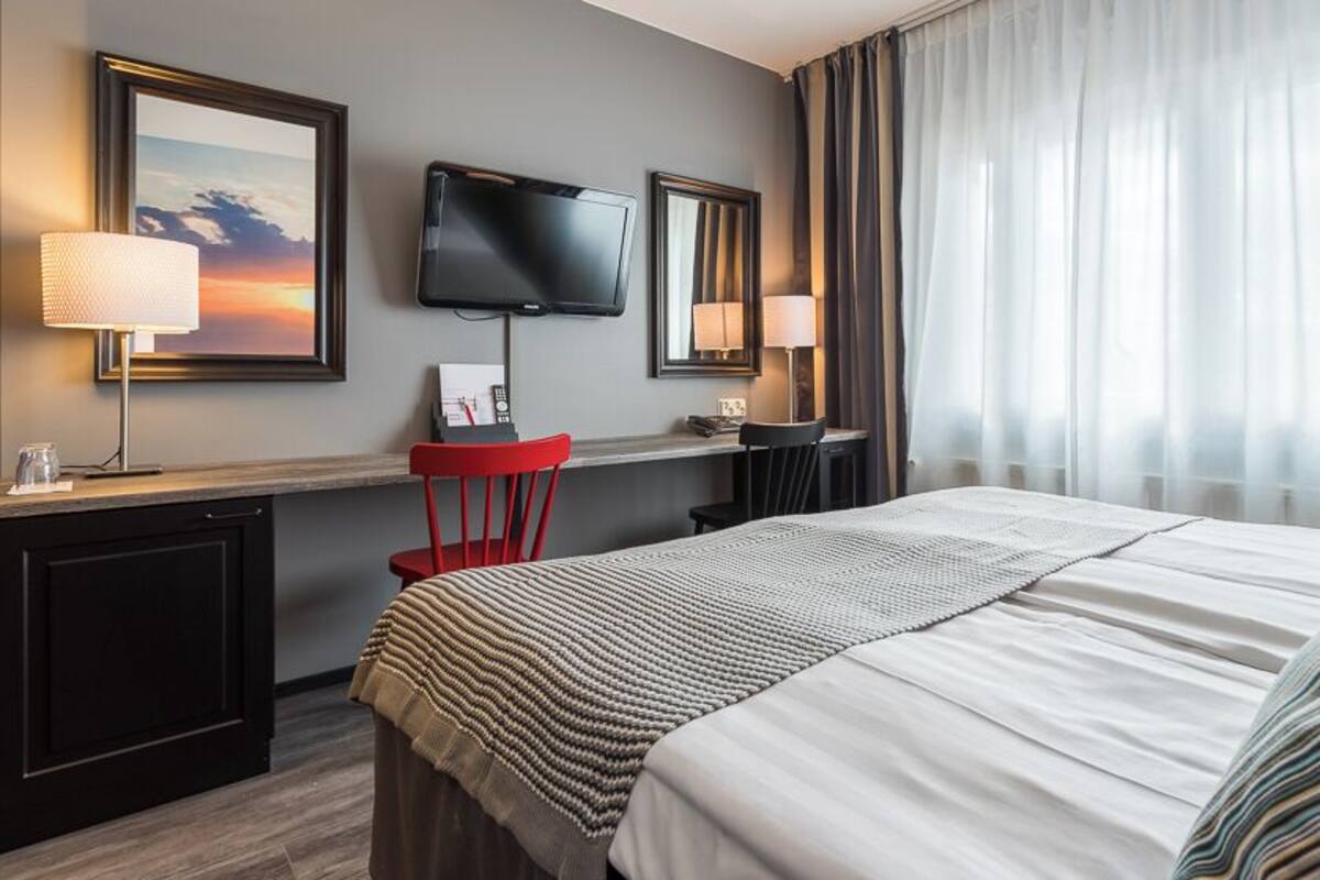 Una camera d'albergo con un letto, una scrivania, una sedia, una televisione e una finestra con tende.