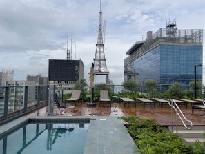 A rooftop pool area with a view of surrounding buildings, including a tall communication tower and a modern glass building. - FV02J - Cozinha - Piscina - Av Paulista (São Paulo)