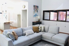 Ein Wohnzimmer mit einem grauen Ecksofa, einem kleinen Beistelltisch und einem Fenster mit Jalousien.