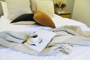 Uma cama bem arrumada com um edredom branco, um travesseiro marrom e um travesseiro preto. Uma xícara de café e um livro estão sobre a cama.