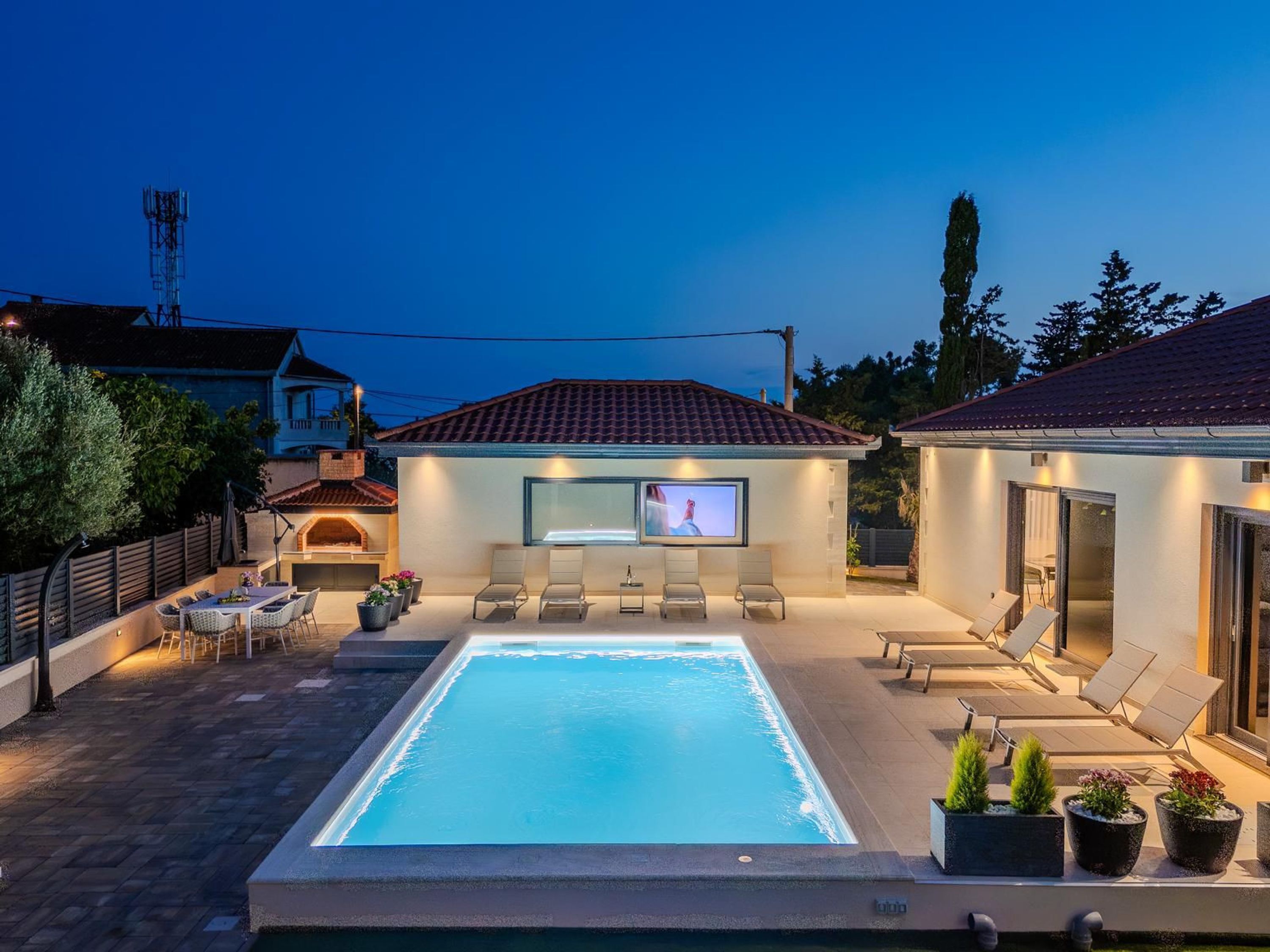 Una piscina con pantalla de televisión, sillas de descanso y zona de barbacoa.