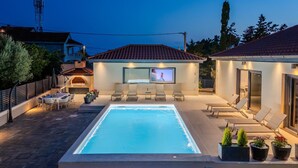 Une piscine équipée d’un écran de télévision, de chaises longues et d’un espace barbecue.